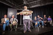 A bearded person stands, holding an enormous fish on a small, all-black stage while seven people sit on chairs behind.