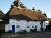 A wide, white-painted building with a prominent thatched roof and two chimneys. There are nine irregularly spaced windows; those at first-floor level are immediately below the roofline and are smaller. A blue door is set in the left-hand side, and a white gate is set next to it in a flint wall. A stumpy, possibly dead tree obscures the rightmost two windows.