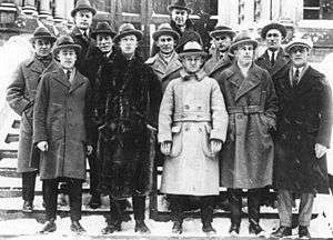 Twelve men pose on the steps in front of a building. They are wearing suits, long jackets and hats.