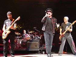 Four men on a stage, all wearing black clothing. Two are playing guitars, one is sitting behind a drum set, and one is holding a microphone up to his mouth. In the background is a crowd of people.