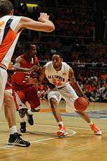 A person wearing a white-colored basketball uniform holding a basketball passing by another person wearing a red basketball uniform in front of another person also wearing white.