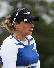 A blonde-haired woman in a blue front shirt with white sleeves and a black hat with sunglasses clasped to her hat backwards and upside down