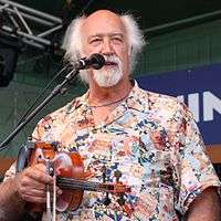 Michael Doucet holding a fiddle and bow in his right hand. He has white hair.