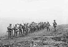 A line of soldiers in battle equipment face another soldier who is addressing them on a gentle slope.  Behind them, smoke or fog obscures the rest of the terrain.