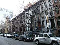 A row of similar three-story townhouses in different colors with stoops on a city street with bare trees and cars parked in front of them. At the left end is a yellow building with a tower.