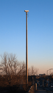 A video camera mounted on a tall cement pole on the side of a roadway. The camera is not pointing at the roadway visible at the bottom-right of the picture, but to the left.