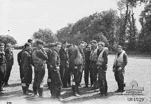 Black-and-white photo of a group of young men wearing military uniforms and life preservers standing in a field edged with trees