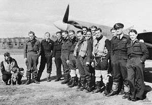 Thirteen men wearing military uniform standing close together in front of a single propeller monoplane.  A fourteenth man wearing military uniform is crouching and patting a dog to the left of the group.