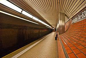 The Lexington Avenue–63rd Street station platform, with a subway track on the left, beige platform tiles, and an orange false wall on the right