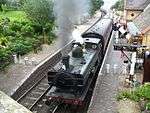 A green pannier tank locomotive is seen from above and to the front as it waits at a station with tow railway tracks and platform. The platform on the left includes a small wooden shelter, benches, lamps, flowers, and, near the end of the platform, three milk churns positioning across the platform to prevent people passing. The platform on the right, where the train is waiting, includes a brick building, more flowers, and again, three milk churns arranged to stop people passing. There is also a signal near the locomotive, and its black and white semaphore arm is pointing downward. The train includes seven coaches in maroon. A plume of grey smoke comes from the locomotive chimney, and a smaller plume of white steam from the safety valve near the cab.