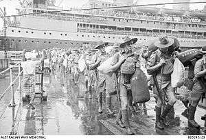 Men in shorts and slouch hats with rifles slung, carrying duffel bags march along a wharf. In the background is a cruise ship.