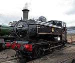 A pannier tank locomotive is seen from the front and to the right and is standing over an ash pit. It is all in black apart from a number of small details. The buffer beam at the front is red. The locomotive number, 9600, is shown in white on a plate on the smokebox door and on a larger plate, edged in white, on the side of the cab. There is a small yellow disc, with the letter C above the number plate. The shed number, 84 E, is shown in white on a small plate near the bottom of the smokebox door. The pannier tank and cab side are lined in red and white, and the British Railways crest, in yellow, white and red, is on the side of the pannier. The letters T Y S are shown in white on the running plate step near the front. The coupling rods are unpainted and are steel grey. Finally, two white lamps are stored next to the black toolbox near the back of the running plate.