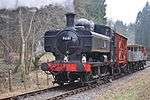 A black pannier tank locomotive is passing along a single track through woodland. The locomotive is pulling a train of a covered red wagon, three grey open wagons, and a red guard's van.