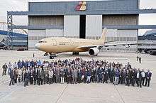 Crowd of people assembled in front of unpainted aircraft. A tall building serves as the backdrop for the photograph