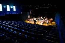  Empty blue seats and a small orchestra on a lighted stage, as seen from the rear of an auditorium