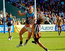 Two young male athletes contest the football. One is wearing a light blue, black and white sleeveless shirt and white shorts, while the other is wearing a brown and gold stripped sleeveless shirt and brown shorts.