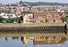 A river scene with wharf and above it a two storey red brick building. The building has decorative brickwork and plenty of windows. Along the facade are teo three storey decorated gable features.