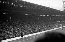 Photograph of a stand full of people. In front of the stand is a field and a goal.