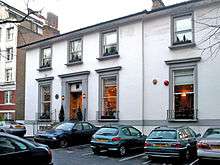 A number of cars are parked in the asphalt car-park of a two-storey white building.  The building appears once to have been detached, but a two-storey extension is apparent, on the right side of the building.  The ground floor contains three large sash windows, each with a small metal railing around the sill.  The first floor contains four shorter sash windows.  The decorative stonework around the windows and main entrance has been painted grey.  Two chimney stacks are visible on the roof, at opposite ends.  The staircase entrance to the building is bordered by metal railings.  To the left of the image, a much larger brick building can be seen in the distance.