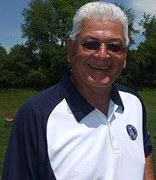 A smiling white tanned man with grey hair, wearing sunglasses and a blue-collared white shirt, outside on a playing field.