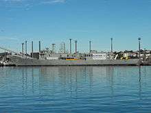 Adelaide tied up alongside at a wharf. The majority of her communications and radar masts have been removed, she carries no weapons, and several large squares have been cut into the ship's hull.