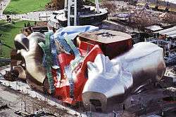 Aerial view of a large city building. The building is composed of oddly shaped, metallic sections. Curved glass and metal pieces run along the face of the building.