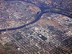 High-angled aerial shot of a developed city, a suburban grid dominates the lower half of the image. A river bisects the city from the left before forking; the first fork continues up and to the right edge of the image; the second curves up and around to finish on the left, enclosing industrial units and other domestic properties.