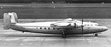 Twin-engined, triple-tailed, high-winged propeller-driven passenger aircraft, in profile on an airport taxiway. Natural metal finish except for airline insignia and a lengthwise stripe along the length of the fuselage.