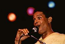 A man with his eyes wide open, holding a microphone placed on a stand. In the background are three stage lights, each a different color