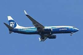 Right side view of an aircraft painted blue on the fuselage in Boeing's corporate livery, with Alaska Airlines' Eskimo face on the tail. The aircraft is airborne in blue skies.