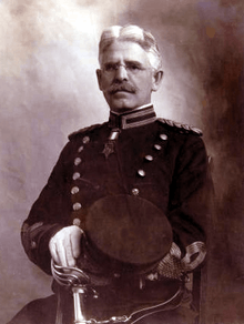 Head and torso of a seated white man with mustache and gray hair wearing a military jacket and white gloves, holding a peaked cap in one hand with the hilt of an ornate sword resting against his leg.