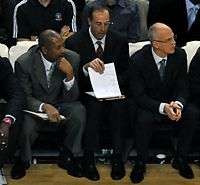 Three men, wearing black suits, white shirts and ties, is sitting in front of a crowd.