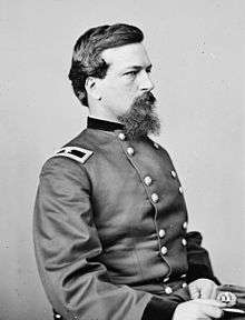 Profile of a dark-haired white man with a mustache and scraggly goatee, wearing a double-breasted military jacket and sitting with a military cap in his lap.