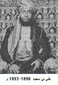 A black-and-white photograph of a man with a dark beard wearing a turban, a dark jacket, and a white shirt, sitting, and looking at the viewer