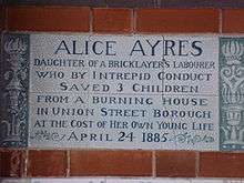 A tablet formed of six standard sized tiles, bordered by green flowers in the style of the Arts and Crafts movement, with an inscription in green capital letters on a white background