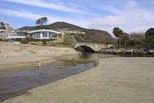 A creek flows out of a gap through a coastal mountain range and flows onto the beach.