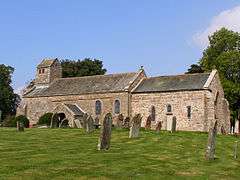 A long, low stone church seen from the south with a west bellcote surmounted by a saddleback roof