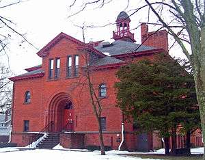 A brick building with an arched entrance, pointed front roof and rounded tower on top viewed slightly from its right. There is snow on the ground.