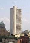 Elevated view of a 35-story skyscraper with a rectangular cross section; the lateral side of the building has a windowless, concrete facade, whereas the anterior side has an almost complete glass facade that is light gray in color. A concrete shaft extends up the anterior side and projects beyond the roofline of the building. An antenna mast extends from the building's roof.