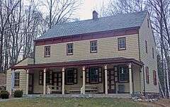 A two-story light yellow house with reddish trim, a pointed roof on the side, a small shed-roofed addition on the left and a porch along the front.