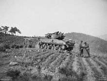 A main battle tank in a cultivated field. To the rear are two soldiers holding a map