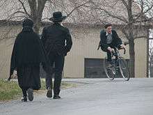 A couple in dark clothes walk along a road, backs to the camera, while a young man on a bicycle heads in the opposite direction.