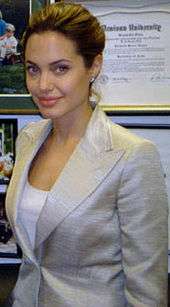 A brown haired woman stands in front of a wall with framed pictures and papers. She wears a light grey jacket over a white top.