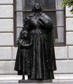 A photograph of a metal statue of a woman standing upright with her head tilted upward and her eyes looking up.  She is dressed in a full dress, and beside her is a young girl who is clinging on to her.