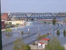 A street with a major amount of water on it due to flooding. A bridge is on the top of the image, and a row of buildings to the left. Sandbags are in front of the buildings