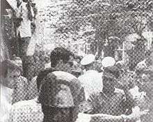 A crowded and narrow street with a lot of people in the vicinity. A tall, Caucasian man with brown hair and sunglasses stands in front of a smaller Asian man with black hair, shielding a shorter man in light shirt with a bloodied face. The smaller Asian man puts an open palm in the air to acknowledge the taller man. A steel-helmeted policeman is in the foreground and others in white caps are in the background.