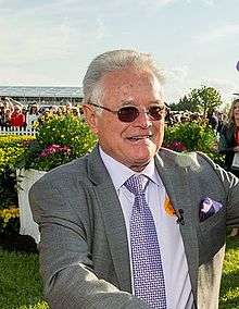 a smiling elderly man in a gray suit, wearing sunglasses