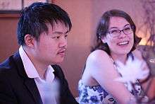 Chu sits foreground left in a black suit besides Blair, background right, in a blue and white floral dress.