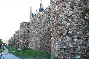 Photo shows a 10-meter high stone wall made from irregular pieces. Round towers are spaced every 20 to 30 meters along the wall. A path runs along the outside of the wall.