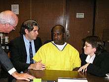 An African-American man in a yellow jumpsuit sits at a desk at the center of the photo, with a white man in his 30s in a business suit on his right, facing him. On the left and right of these two figures, respectively, are a grey-haired man in a suit, standing, and a young, dark-haired woman, crouching, both wearing glasses.